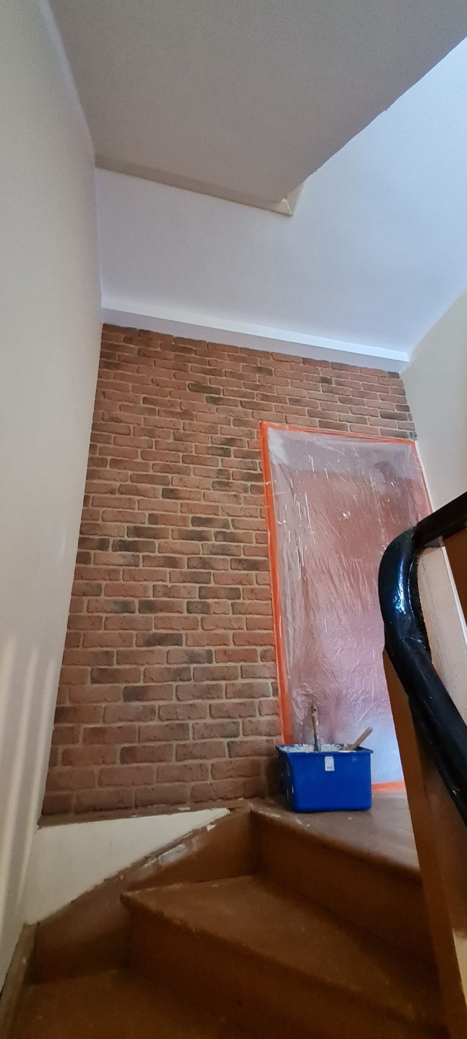 Klatka schodowa w trakcie zabezpieczania przed malowaniem, ściana z cegły dekoracyjnej, zabezpieczone folią drzwi, wiadro z farbą na schodach.