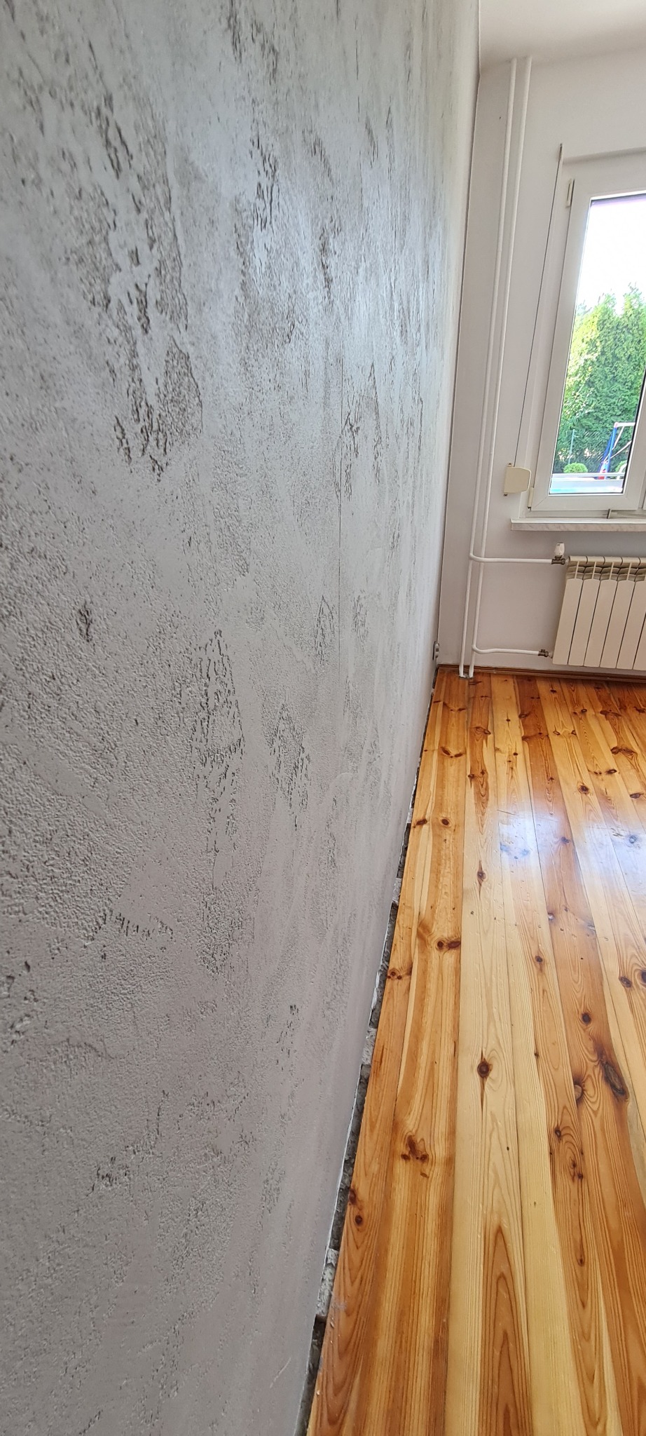 Ściana z efektem betonu, z widoczną fakturą i nierównościami, sąsiadująca z parkietem i oknem z widokiem na zieleń.