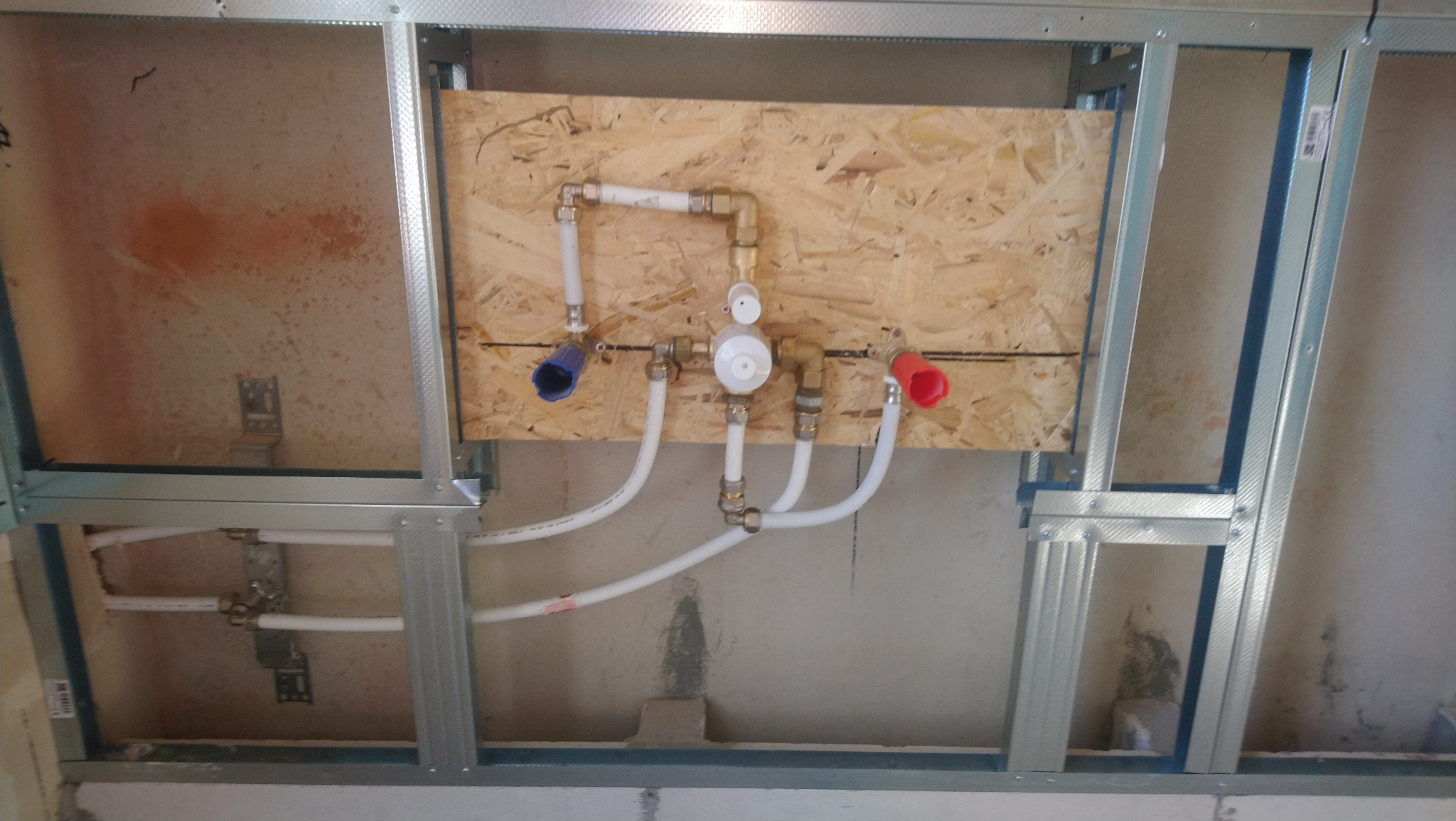 Instalacja wodna z zaworami na płycie OSB, zamontowana w metalowej konstrukcji stelaża na tle surowej ściany w trakcie prac wykończeniowych.