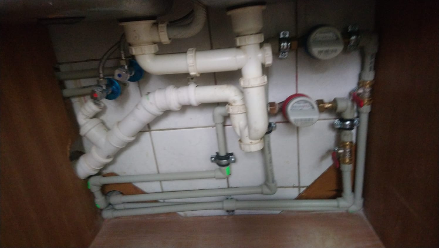 Złożony system rur kanalizacyjnych i wodnych pod zlewem, z widocznymi zaworami, licznikami i elementami mocującymi, na tle płytek ściennych i fragmentu szafki.