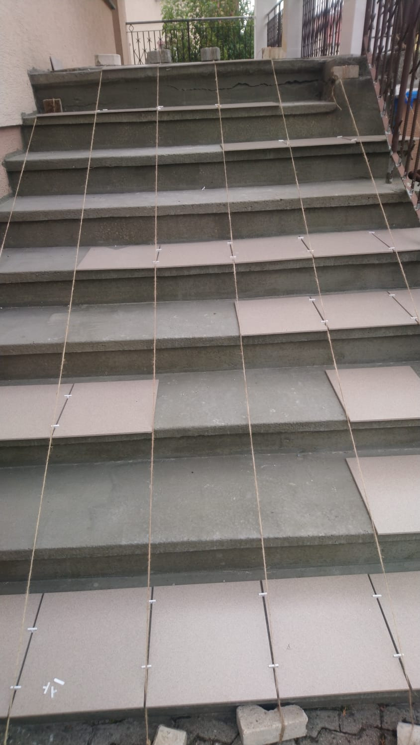 Kładzenie płytek na schodach zewnętrznych, z użyciem sznurków do wyznaczania linii, widoczne białe krzyżyki dystansowe i zaprawa.