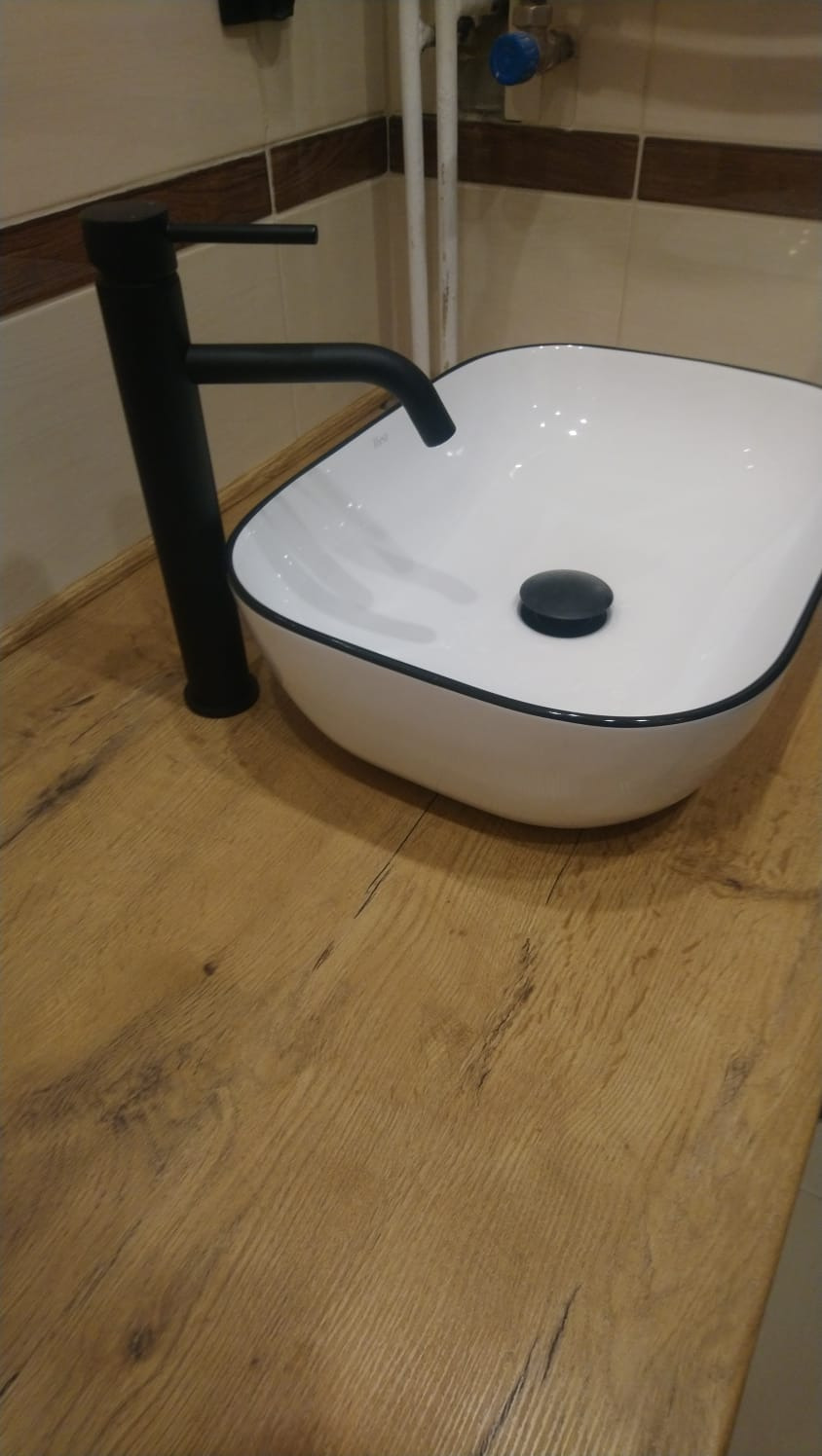 Biała, owalna umywalka nablatowa z czarnym obramowaniem i czarną baterią na blacie z imitacji drewna, widoczne rury instalacyjne w tle.