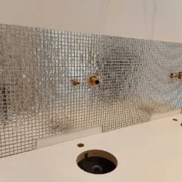 Ściana wyłożona srebrną mozaiką w łazience, z widocznymi otworami na baterie i przygotowanym blatem pod umywalki.