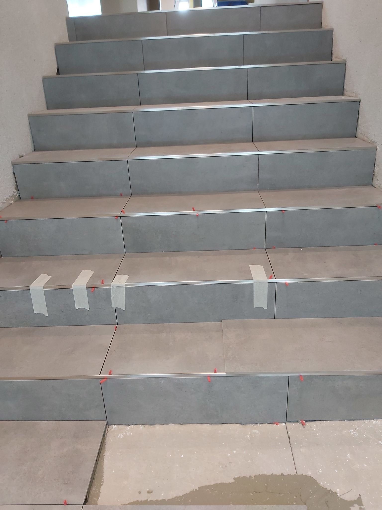 Schody w trakcie wykańczania płytkami ceramicznymi w odcieniach szarości, zabezpieczone taśmą malarską i poziomowane czerwonymi krzyżykami dystansowymi.