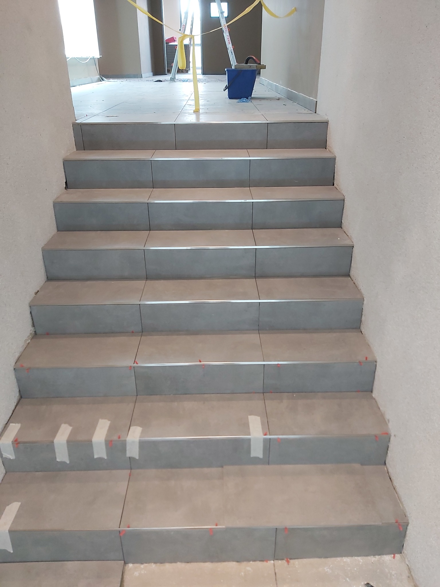 Schody w trakcie układania płytek, widoczne krzyżyki dystansowe i taśmy maskujące, w tle drabina i wiadro.