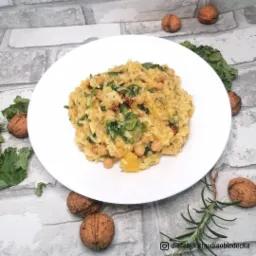 Risotto z ciecierzycą, jarmużem i suszonymi pomidorami na białym talerzu, otoczone orzechami włoskimi i gałązką rozmarynu na tle szarej ściany imitującej cegłę.