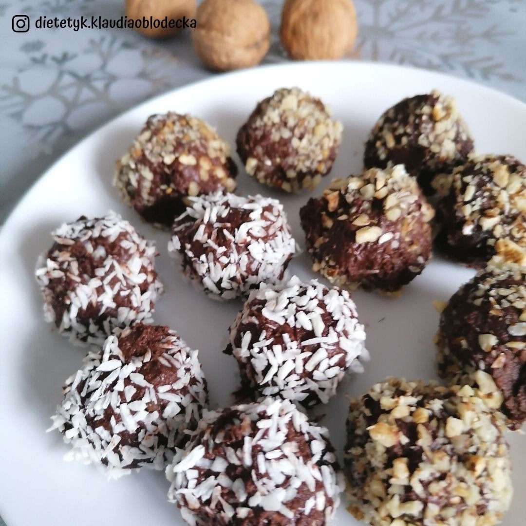 Talerz z czekoladowymi kulkami posypanymi wiórkami kokosowymi i posiekanymi orzechami, w tle dwa orzechy włoskie.