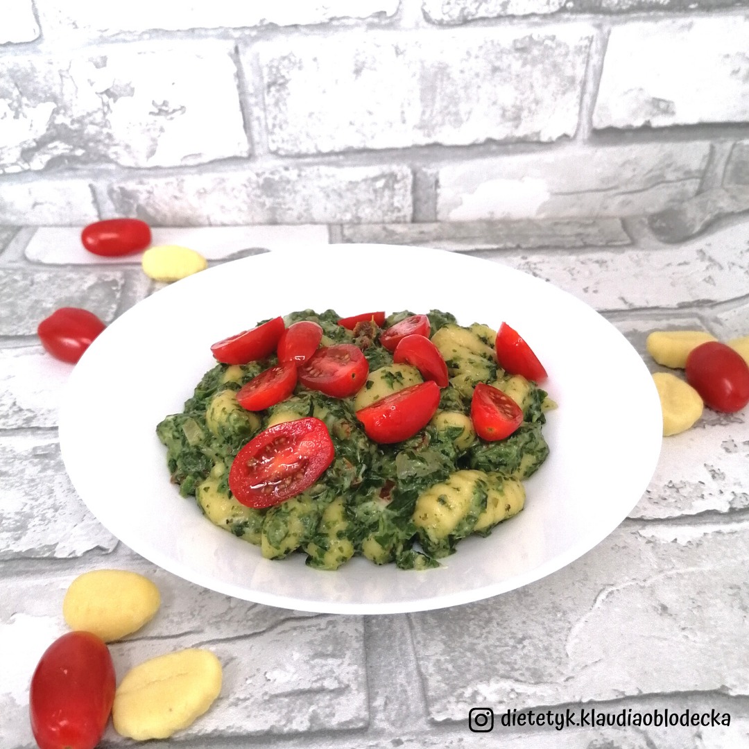 Gnocchi ze szpinakiem i pomidorkami cherry na białym talerzu, tło imitujące cegłę, kilka surowych gnocchi obok, instagramowy podpis w rogu.