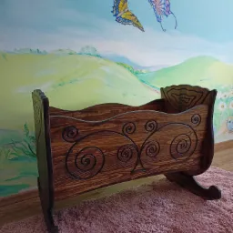 Drewniana kołyska z ciemnego drewna, zdobiona spiralnymi ornamentami, na tle ściany z ręcznie malowanym pejzażem dziecięcym.
