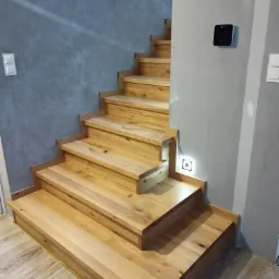 Jasne, drewniane schody z podświetleniem LED w ścianie, prowadzące do góry, z widoczną fakturą drewna i szarym tłem ścian.