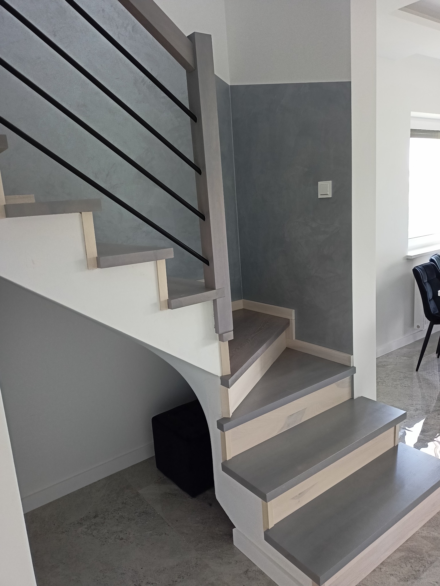 Nowoczesne schody z szarymi stopniami i czarnymi, poziomymi balustradami, widoczne z dołu, z małą pufą pod najniższym biegiem.