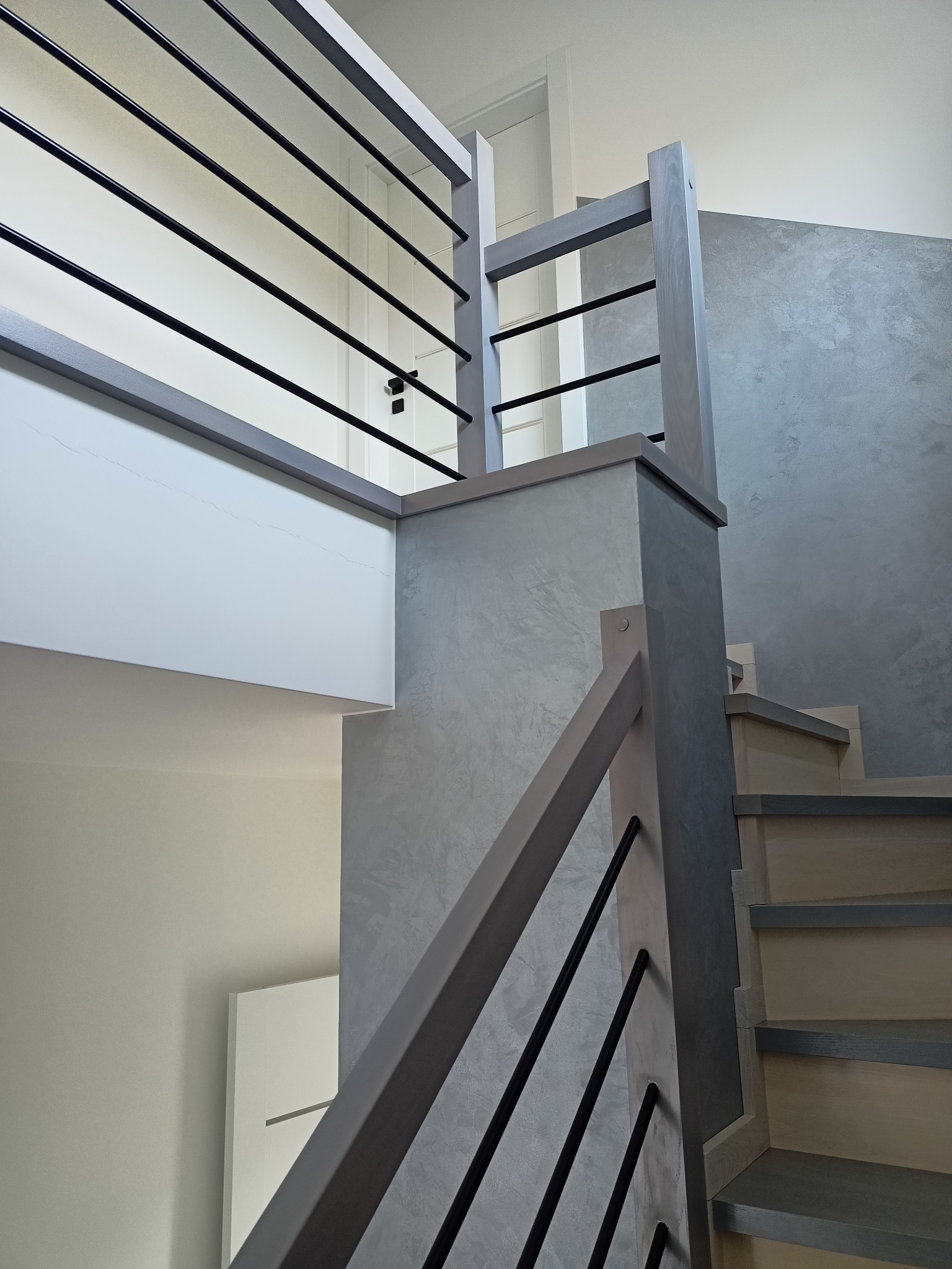 Nowoczesne schody z drewnianymi stopniami i szarą balustradą z czarnymi, poziomymi prętami, na tle ściany z efektem betonu.