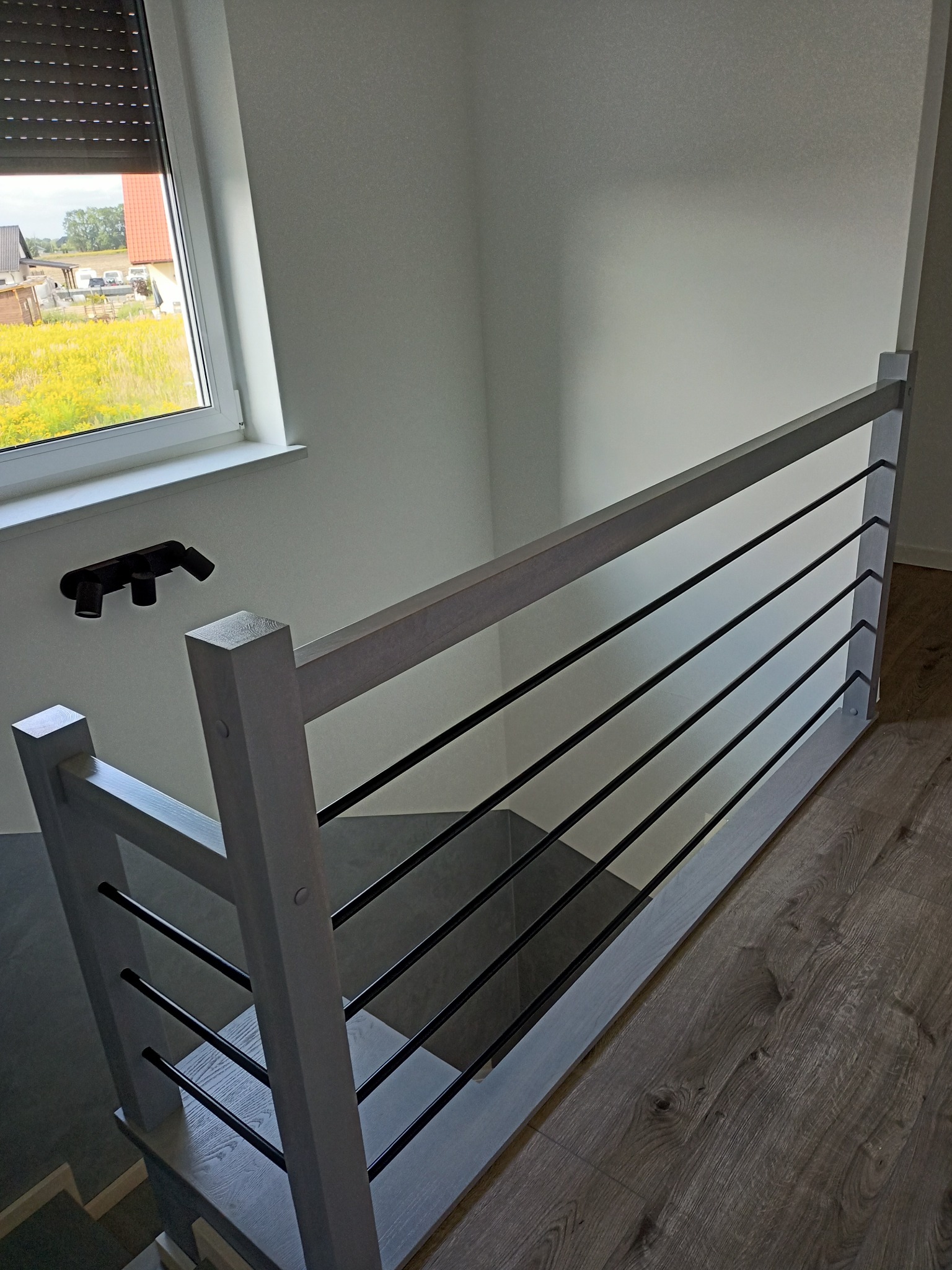 Nowoczesne schody z jasnoszarymi drewnianymi słupkami i czarnymi, poziomymi metalowymi prętami, widok z góry na dół, z oknem w tle i lampą sufitową.