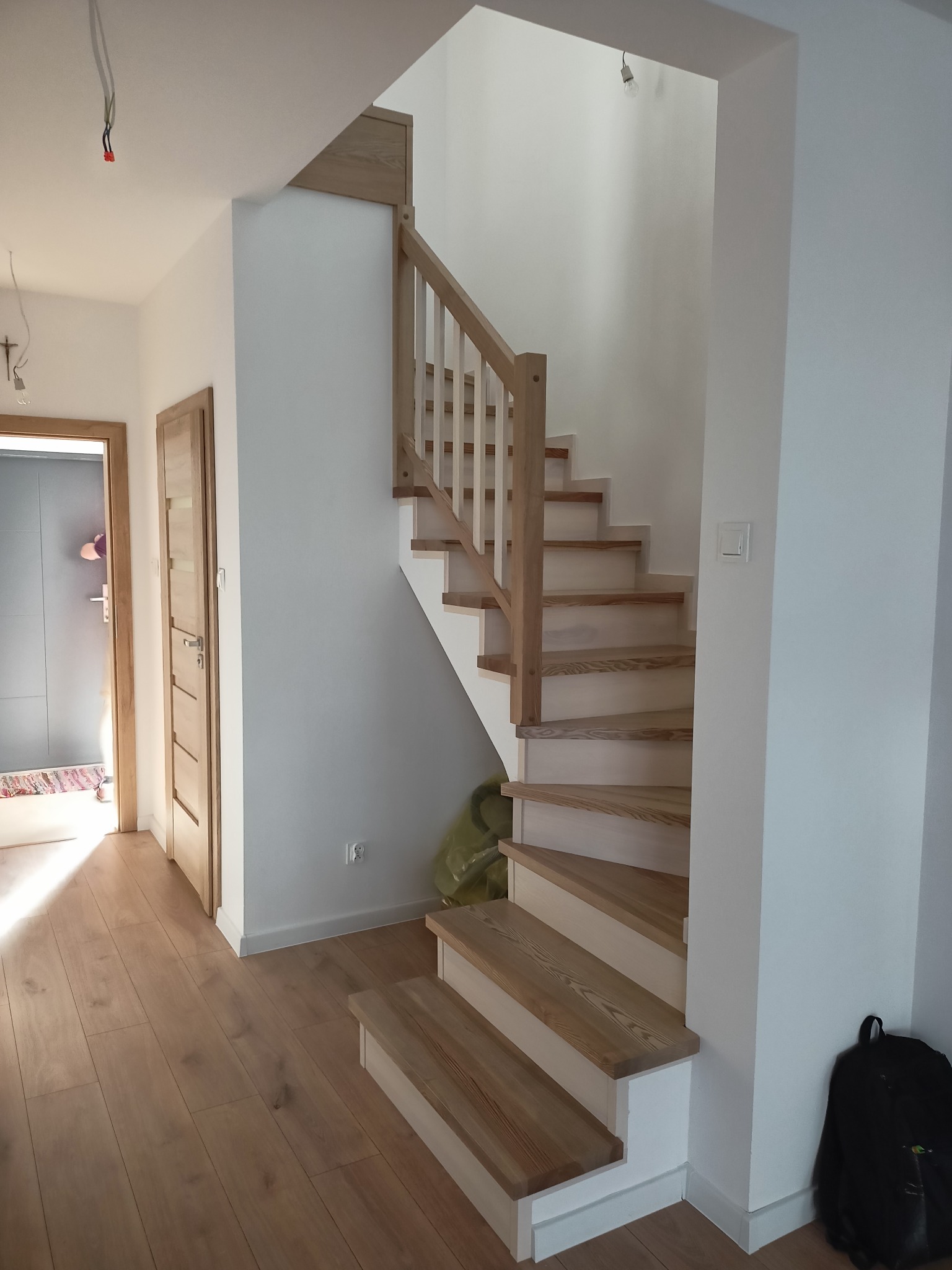 Jasne, drewniane schody z białymi podstopnicami i drewnianą balustradą w nowym domu, widok z dołu na pierwsze stopnie, z plecakiem opartym o ścianę.