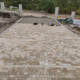 Fragment nowej, beżowo-szarej nawierzchni z kostki brukowej w Szczytnie, z widocznymi kolumnami bramy wjazdowej i przygotowanym podłożem pod dalszą część podjazdu.