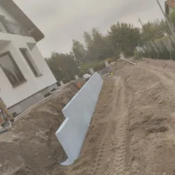 Wkopane w ziemię prefabrykowane betonowe elementy tworzące ścianę oporową, widoczne ślady opon ciężkiego sprzętu, budynek mieszkalny w tle.