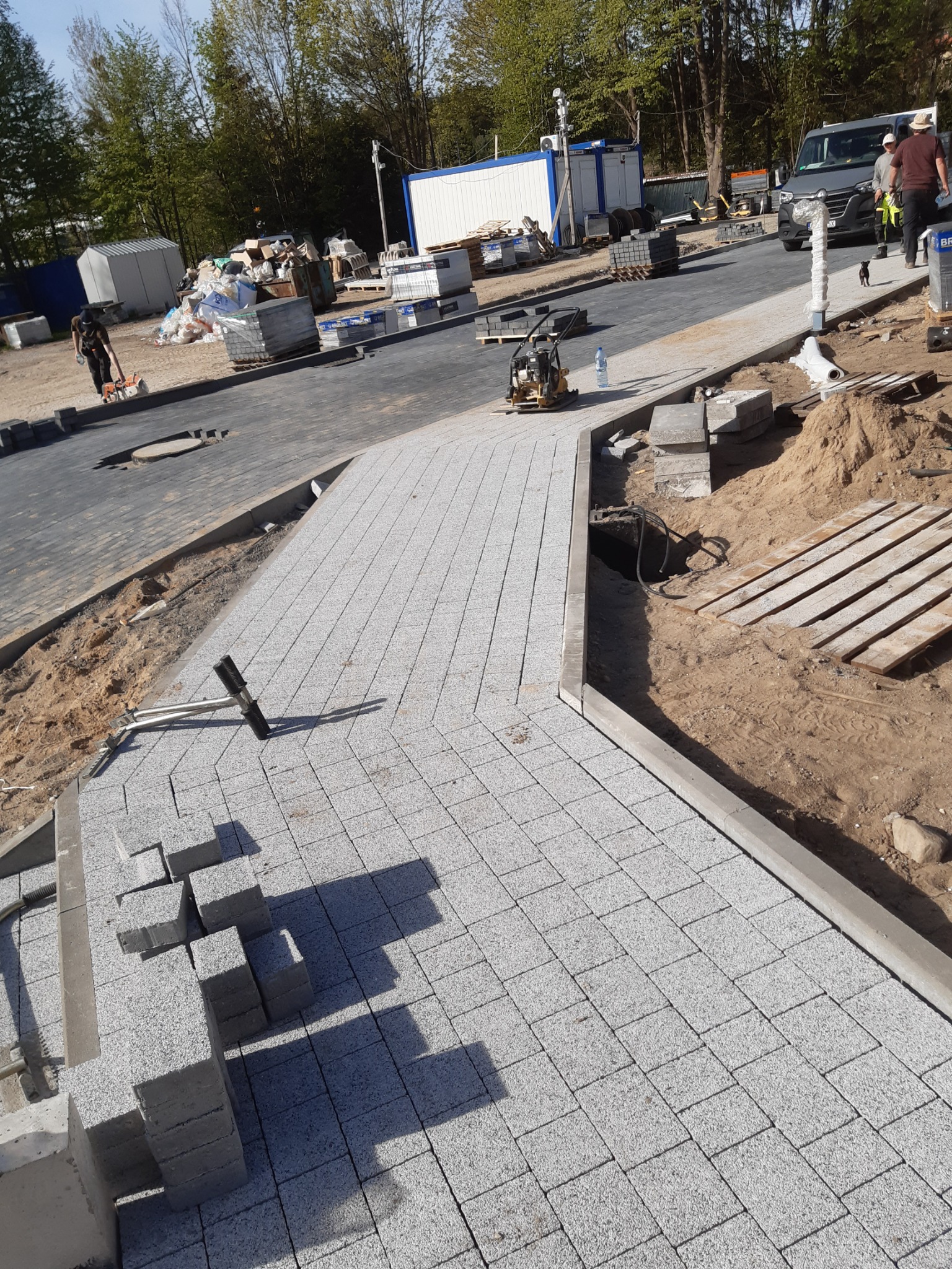 Świeżo ułożona, jasnoszara kostka brukowa na ścieżce, widoczne narzędzia, palety i maszyna do ubijania, w tle budynki kontenerowe i drzewa.