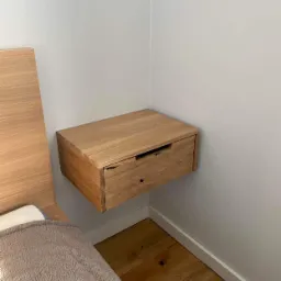 Dębowa szafka nocna wisząca, z widocznym usłojeniem drewna, zamontowana przy łóżku z dębowym wezgłowiem, na tle białej ściany i drewnianej podłogi.