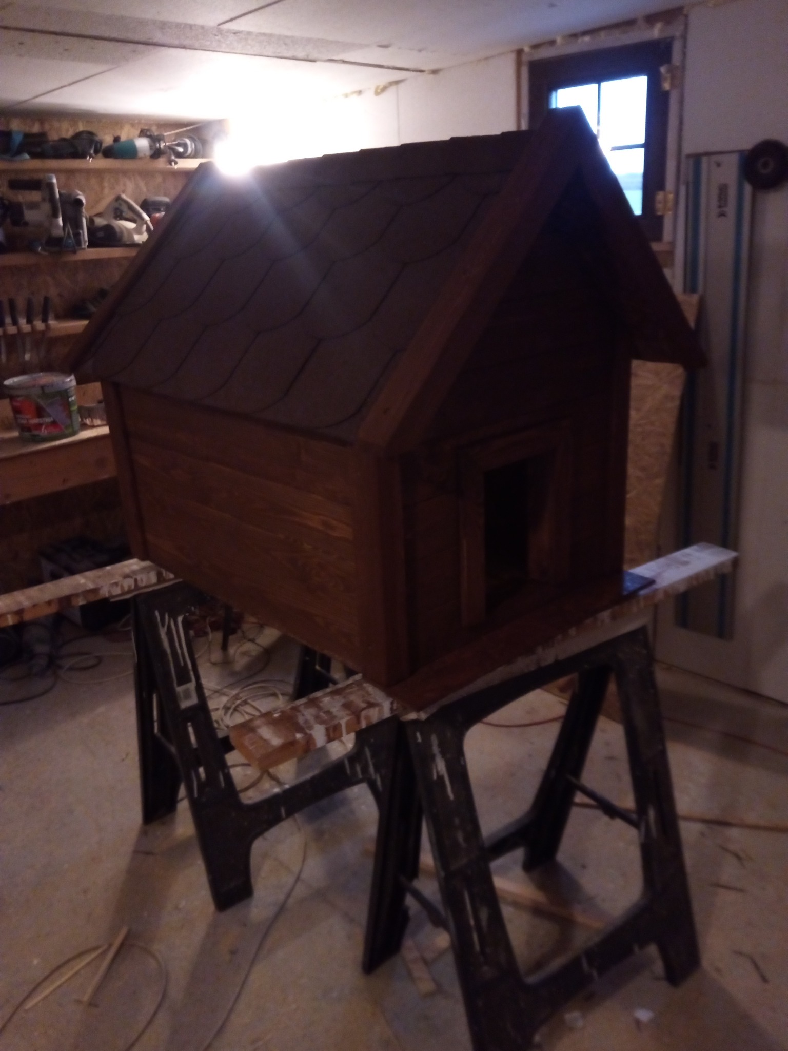 Drewniana buda dla psa z dwuspadowym dachem, umieszczona na kozłach stolarskich w warsztacie, widoczne narzędzia i materiały na półkach w tle.