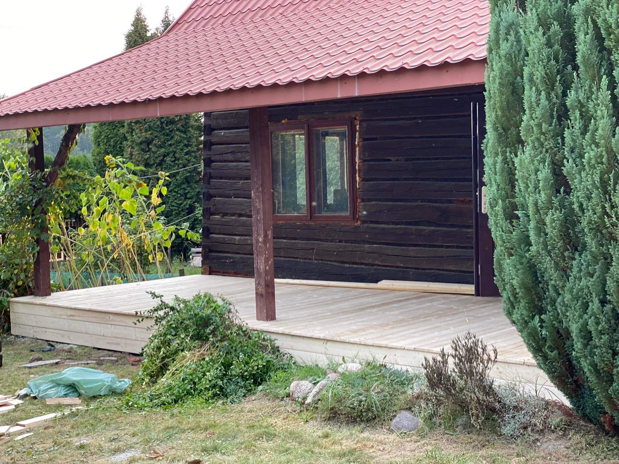 Nowo wybudowany jasny taras przy drewnianym domku z czerwonym dachem, widoczne rośliny, pozostałości po budowie i zielony worek na trawie.