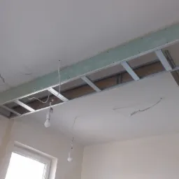 Konstrukcja podwieszanego sufitu z płyt karton-gips, widoczne profile metalowe, przewody elektryczne i żarówki tymczasowe, ściany w stanie surowym.