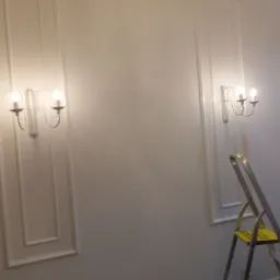 Ściana z dekoracyjnymi panelami i kinkietami w stylu retro; w rogu widoczna drabina malarska z żółtym stopniem.