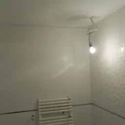 Łazienka w trakcie prac wykończeniowych: białe ściany, dekoracyjne płytki 3D na jednej ścianie, grzejnik drabinkowy i wiszące żarówki na kablach.