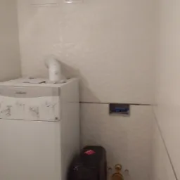 Biały piec gazowy Vaillant z podłączonym kominem w łazience wykończonej białymi płytkami z delikatnym wzorem. Widoczne przyłącza wodne i odpływowe oraz czarny zbiornik. Na ścianie kratka wentylacyjna.