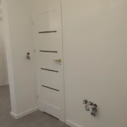 Białe drzwi wewnętrzne z poziomymi, ciemnymi wstawkami, zamontowane w nowym, jasnym pomieszczeniu z widocznymi instalacjami elektrycznymi i hydraulicznymi na ścianie, sugerujące etap prac...