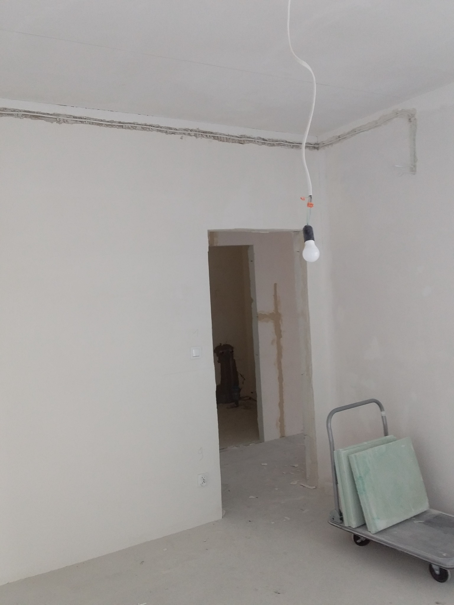 Wnętrze w trakcie prac wykończeniowych: gołe ściany, wystające przewody elektryczne, wisząca żarówka i wózek z płytami gipsowymi.
