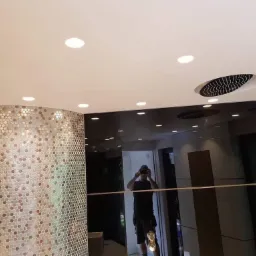 Wnętrze łazienki z kolumną wykończoną mozaiką w odcieniach brązu i srebra, podwieszanym sufitem z oświetleniem punktowym oraz lustrzanymi panelami ściennymi, w których odbija się fotografujący...