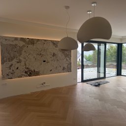 AD-BUD - Salon po remoncie: parkiet w jodełkę, ściana z kamienia z podświetleniem LED, nowoczesne lampy wiszące. Za oknem widoczny plac budowy.