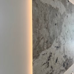 AD-BUD - Ściana z dekoracyjną płytą imitującą marmur, podświetlona pionową listwą LED. Widoczne gniazdka elektryczne w dolnej części ściany.