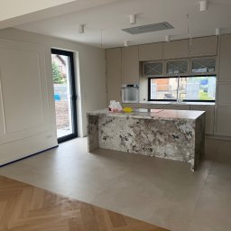 AD-BUD - Jasna kuchnia z wyspą z kamienia i parkietem ułożonym w jodełkę. Widoczne wykończenia ścian i zabudowa kuchenna w odcieniach beżu. Oświetlenie sufitowe punktowe.