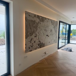 AD-BUD - Wykończona ściana z marmurową płytą podświetloną LED, widoczne gniazdka elektryczne i parkiet w jodełkę, duże okna przesuwne z widokiem na zewnątrz.
