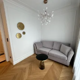 Eleganckie wnętrze: szara sofa, stolik z czarnym blatem, parkiet w jodełkę i designerska lampa wisząca. Białe ściany zdobią złote, okrągłe dekoracje.