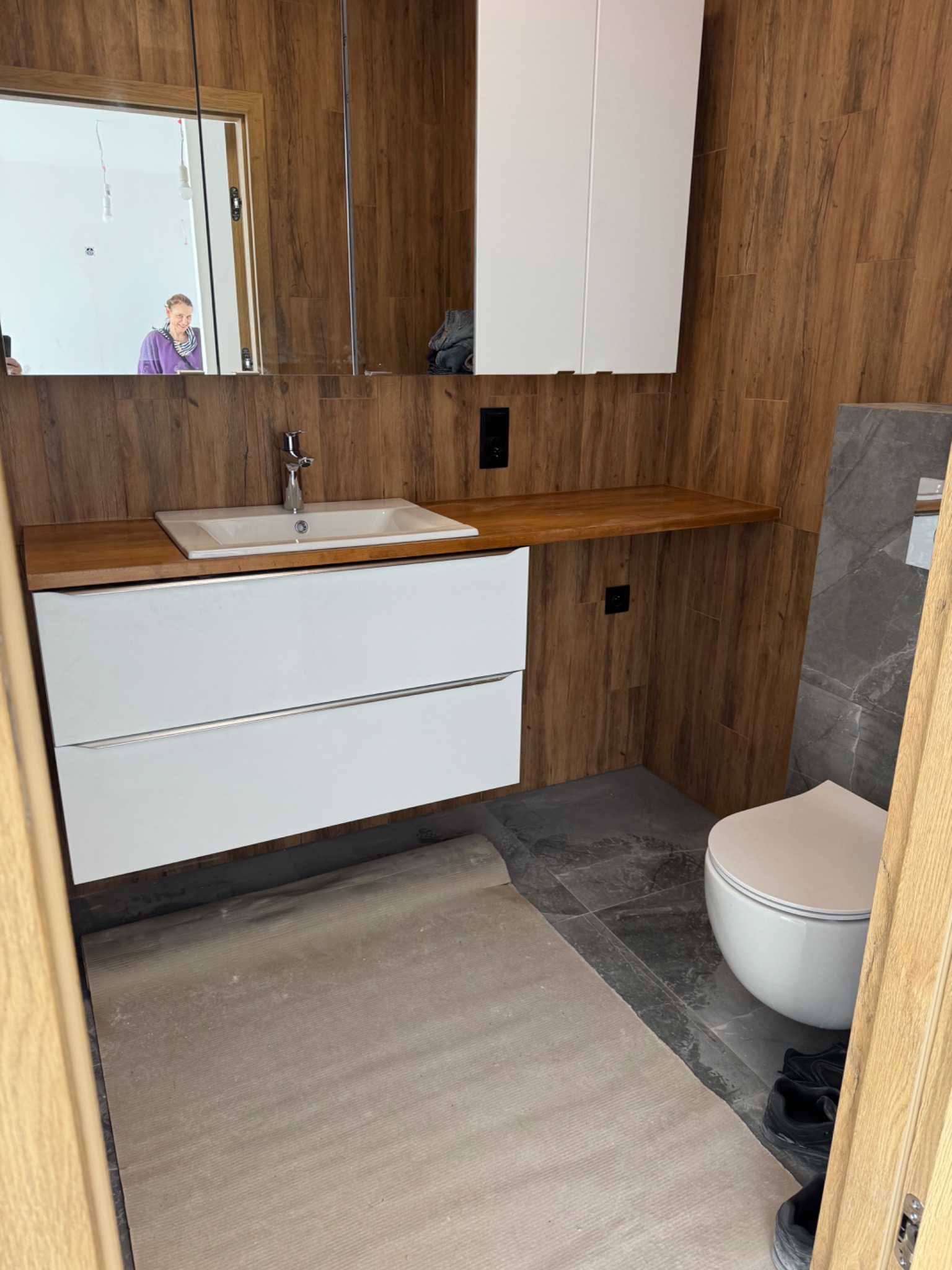 Wykończona łazienka z drewnopodobnymi płytkami, białą szafką pod umywalką i wiszącą toaletą. W lustrze odbija się osoba.