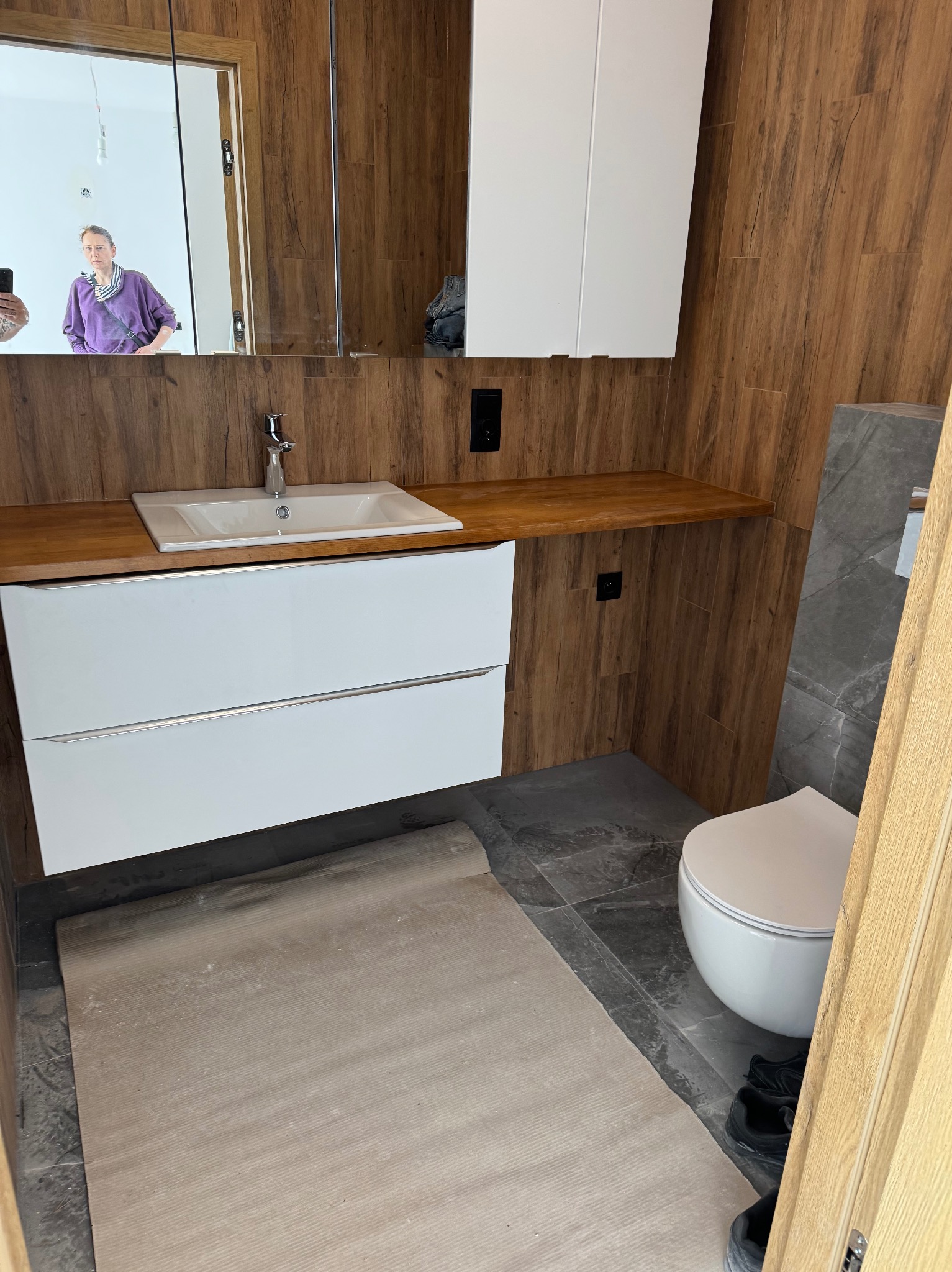 Nowoczesna aranżacja łazienki w Somoninie: drewnopodobne płytki ścienne, biała szafka z umywalką i szara podłoga. Widoczne zabezpieczenie podłogi na czas prac.