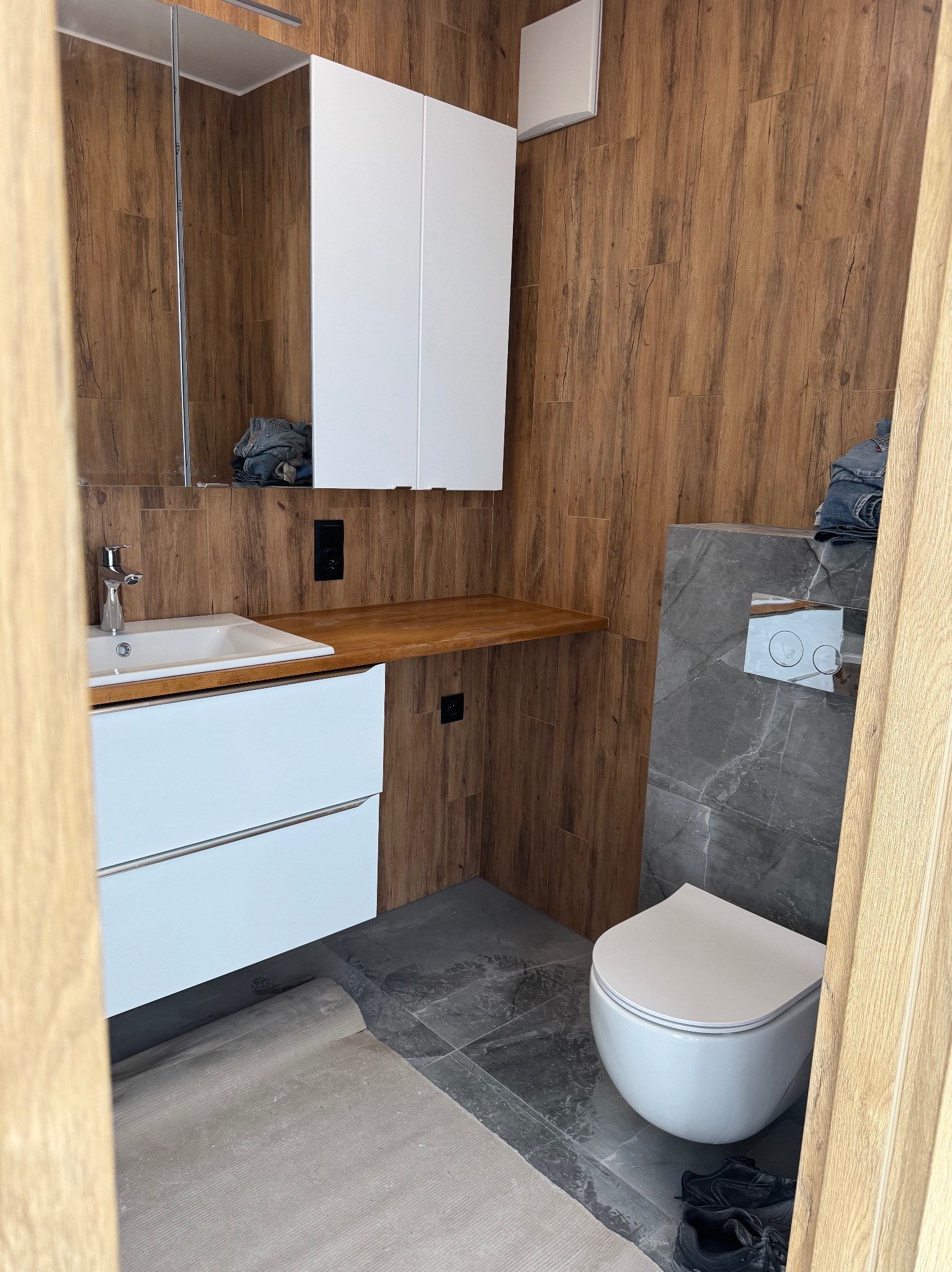 Nowoczesna łazienka po remoncie: drewnopodobne płytki ścienne, biała armatura (umywalka, toaleta), szara podłoga i zabudowa. Widok zza uchylonych drzwi.