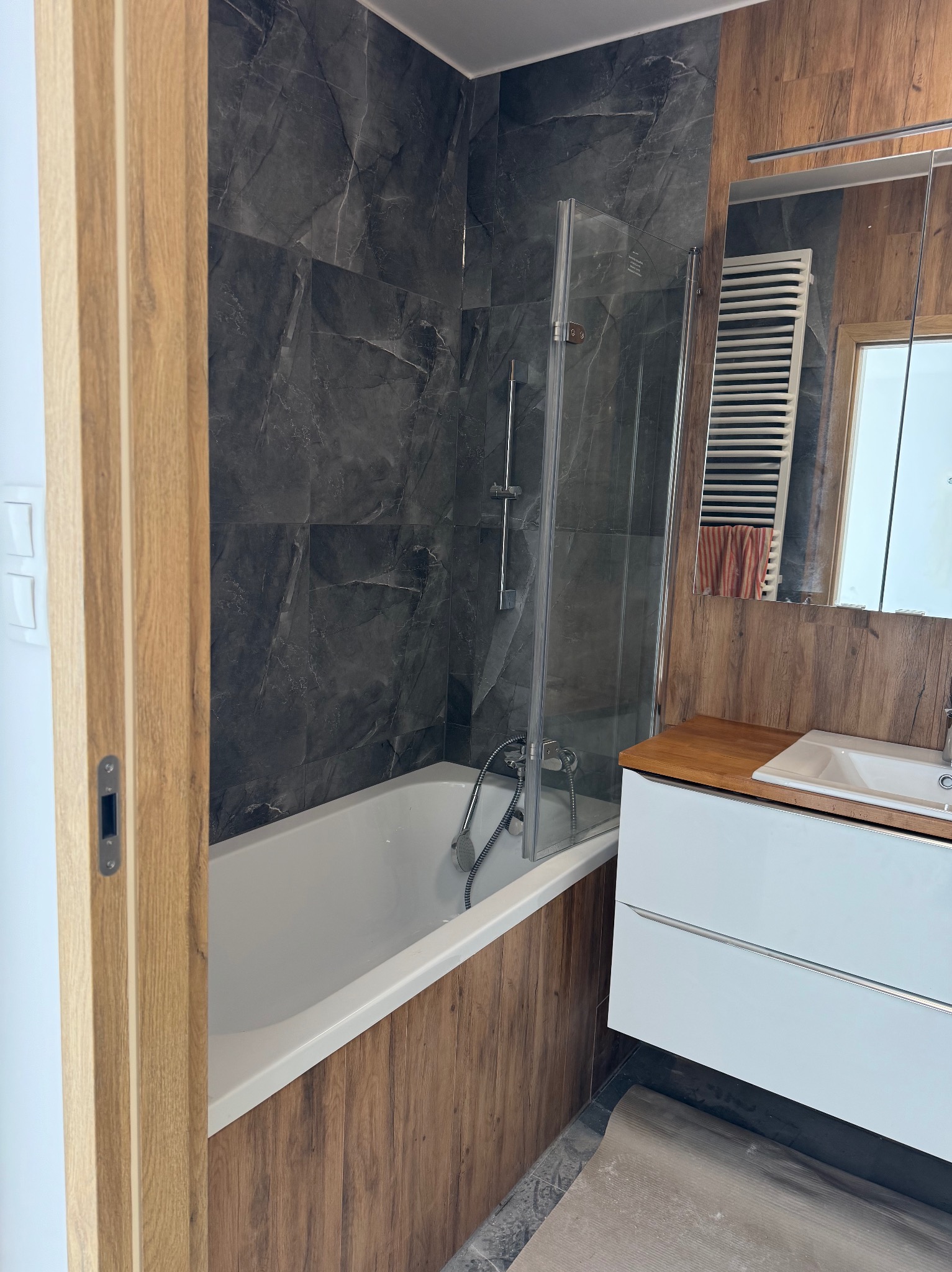 Wykończona łazienka z wanną obudowaną drewnopodobnymi panelami, szklanym parawanem, umywalką z szafką i grzejnikiem drabinkowym w lustrze. Płytki imitujące marmur.