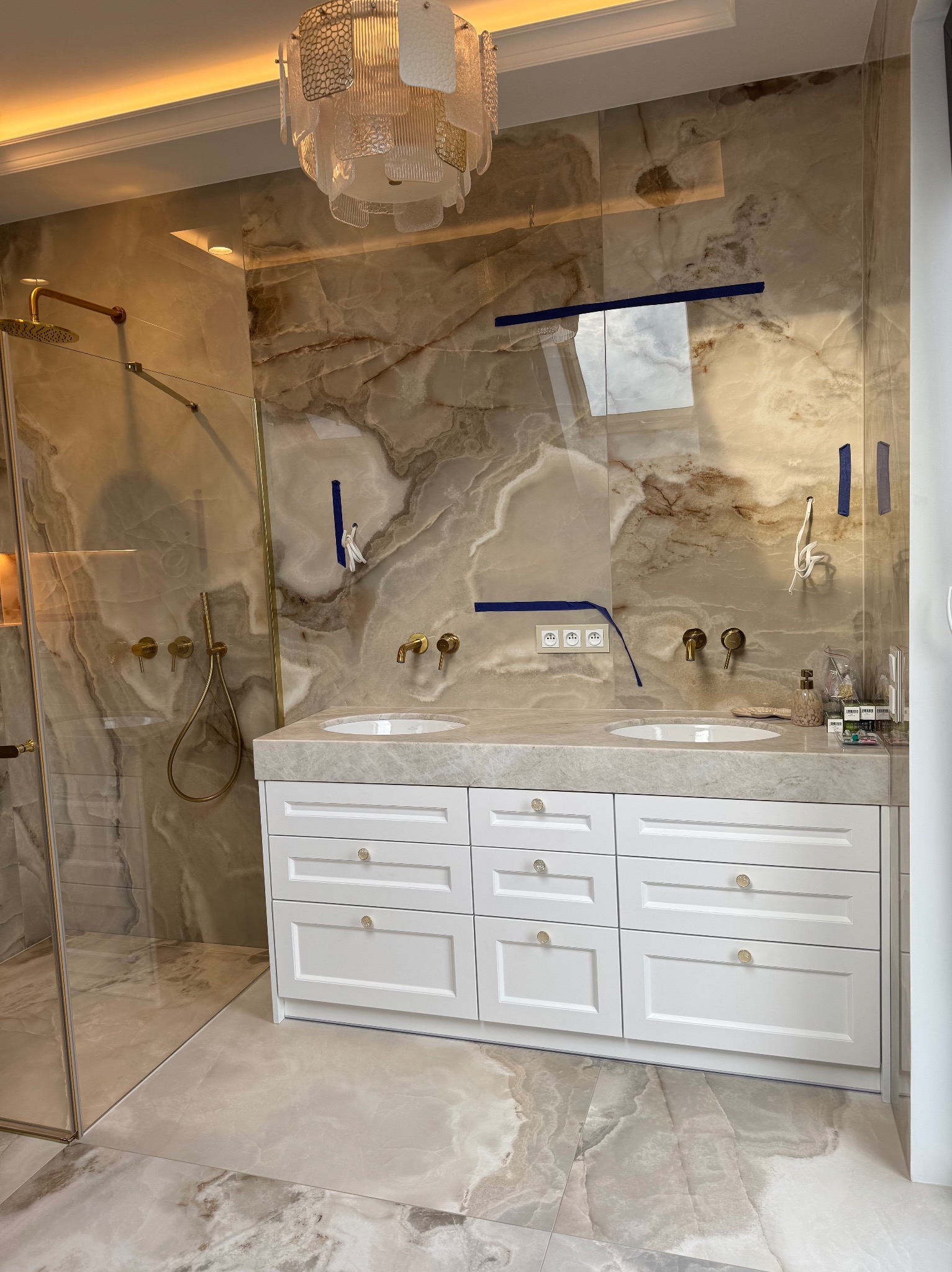 Luksusowa łazienka z marmurowymi ścianami, złotą armaturą i białą szafką pod umywalką. Widoczna kabina prysznicowa ze szklaną ścianką i dekoracyjny żyrandol.