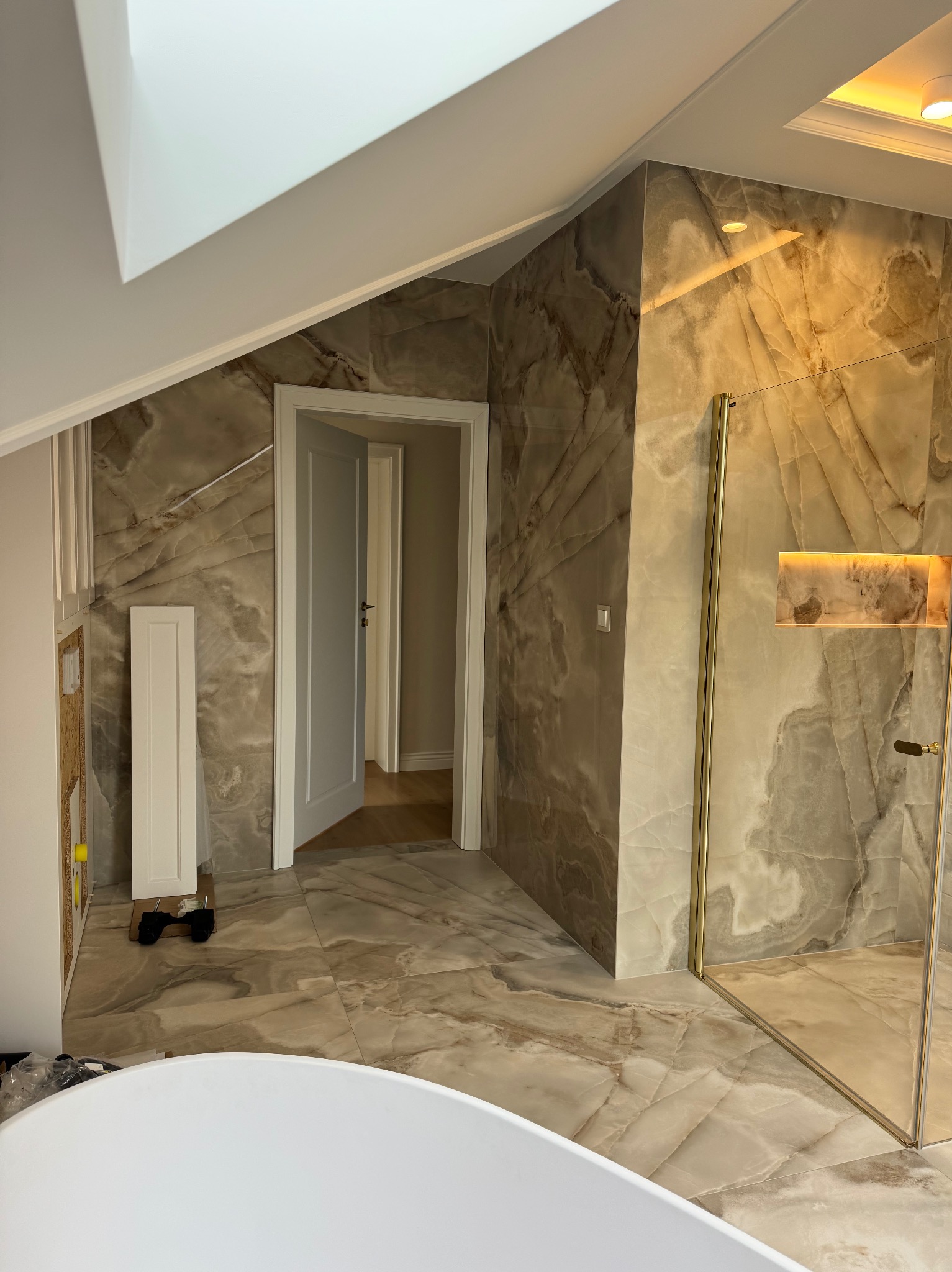 Wykończona łazienka na poddaszu z marmurowymi płytkami, kabiną prysznicową ze złotymi ramami i białymi drzwiami w tle, widok zza wanny.