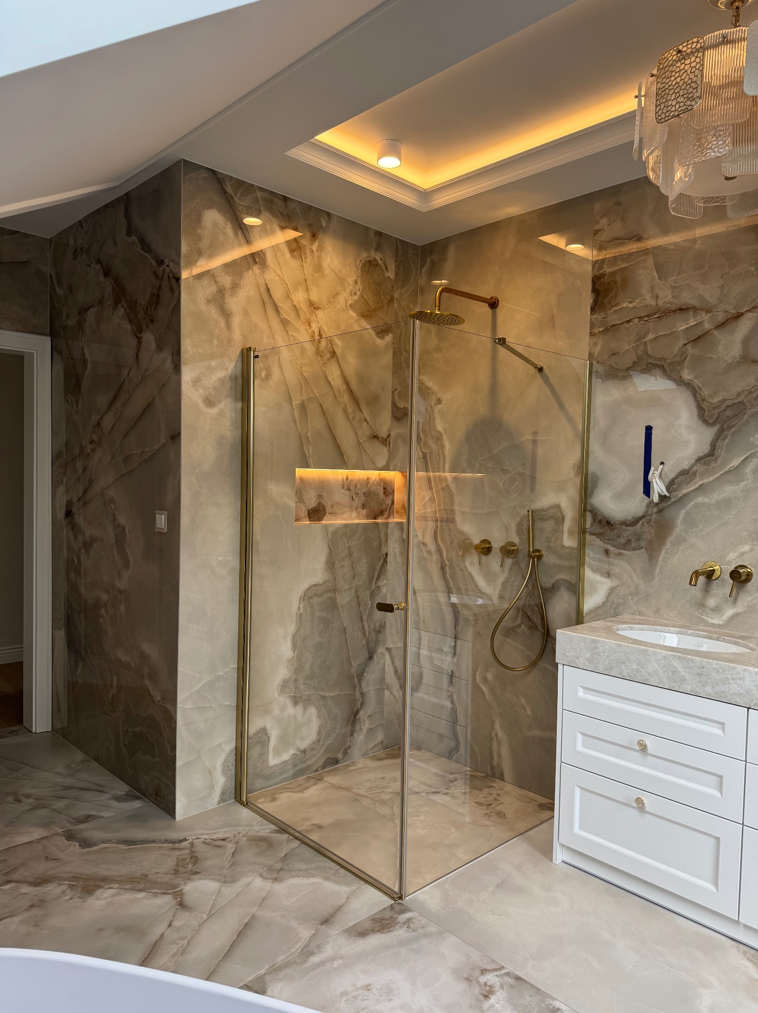 Luksusowa łazienka z kabiną prysznicową w złotej ramie, marmurowe płytki na ścianach i podłodze, biała szafka umywalkowa, designerskie oświetlenie.