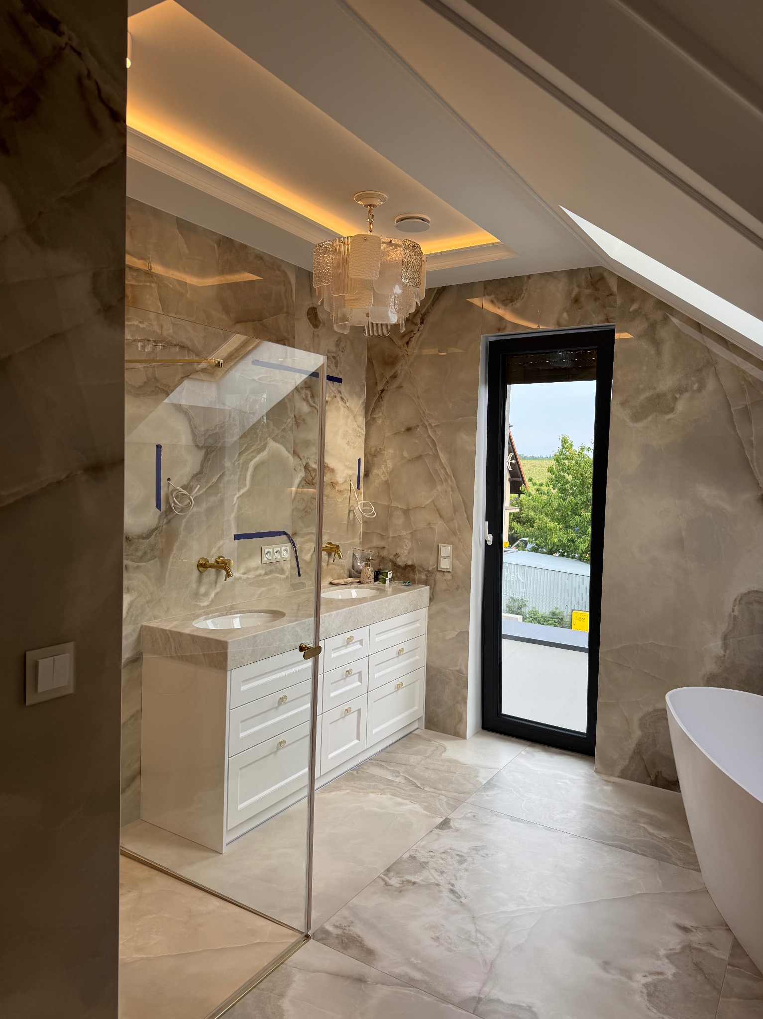 Stylowa łazienka na poddaszu z marmurowymi ścianami, kabiną prysznicową ze złotymi detalami, białą szafką z umywalkami i nowoczesnym oświetleniem.