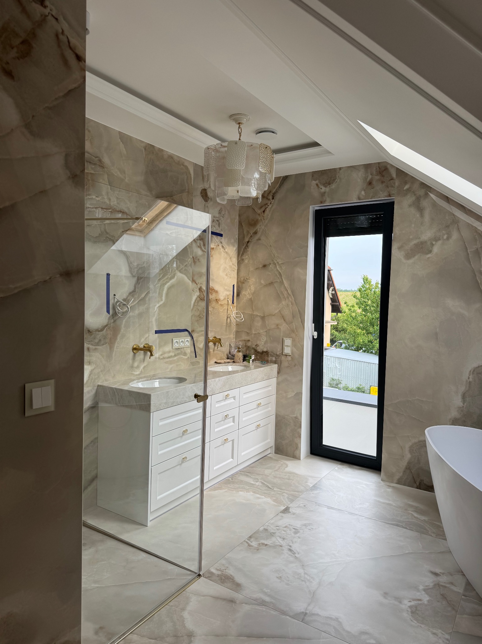 Elegancka łazienka na poddaszu z marmurowymi ścianami, białą zabudową umywalki, kabiną prysznicową ze szkła i oknem z widokiem na zieleń.