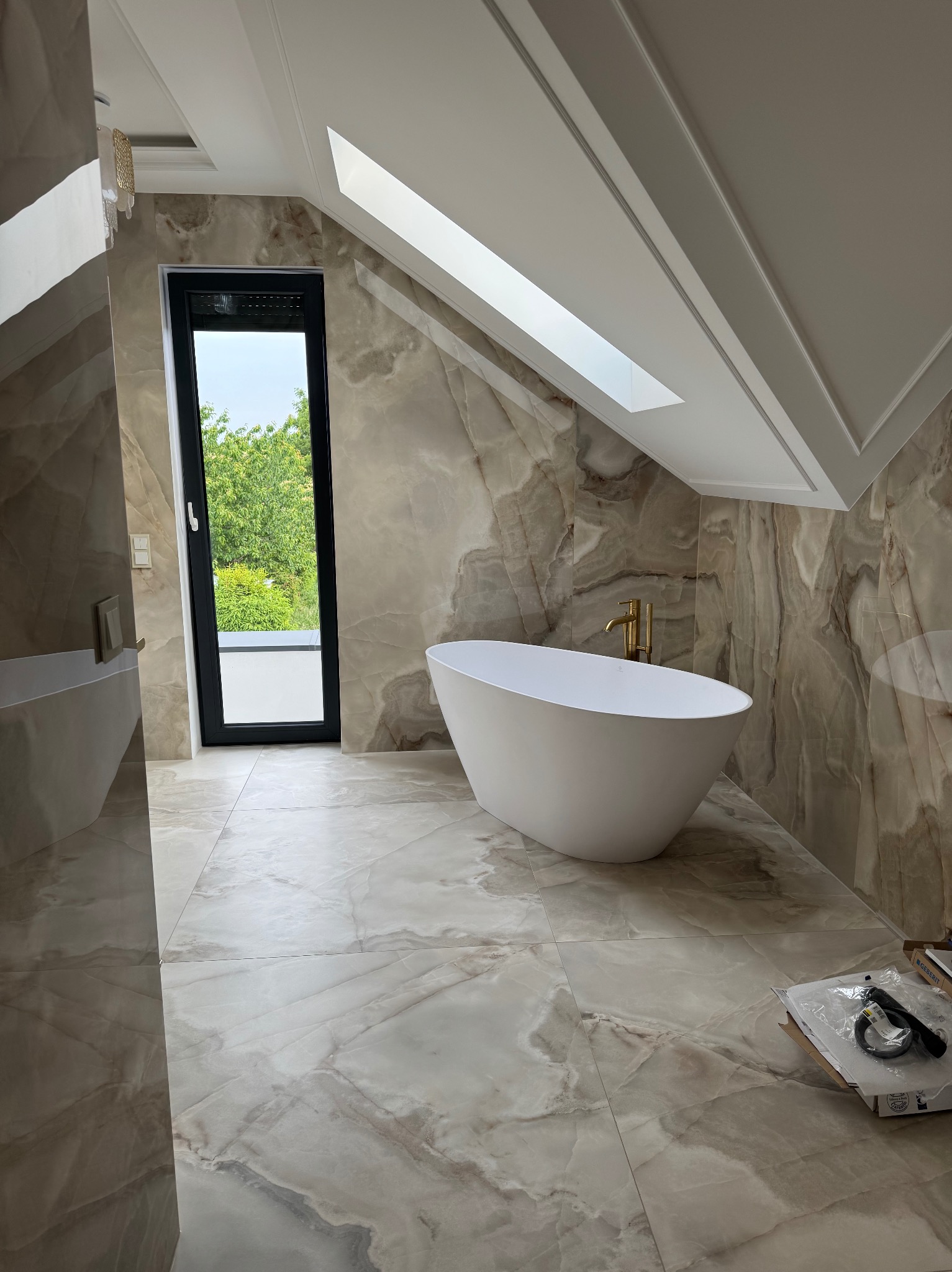 Elegancka łazienka na poddaszu z wolnostojącą wanną, marmurowymi płytkami i oknem dachowym. Widoczne narzędzia sugerują prace wykończeniowe.