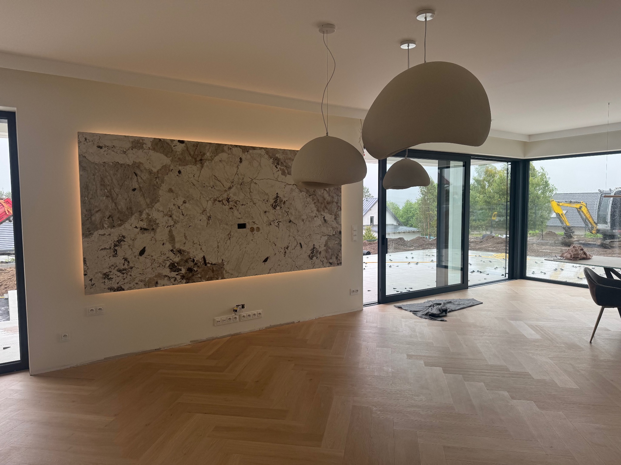 Salon po remoncie: parkiet w jodełkę, ściana z kamienia z podświetleniem LED, nowoczesne lampy wiszące. Za oknem widoczny plac budowy.
