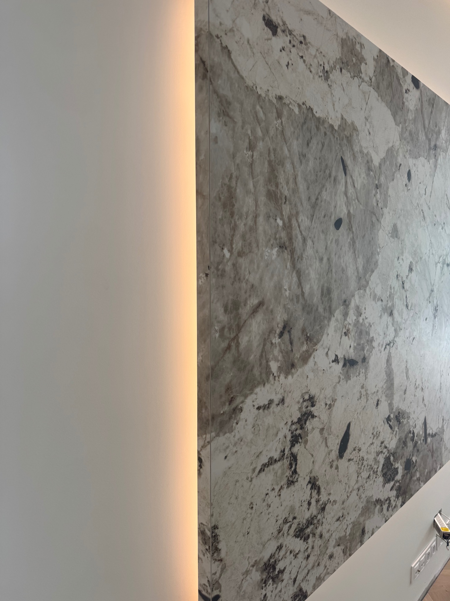 Ściana z dekoracyjną płytą imitującą marmur, podświetlona pionową listwą LED. Widoczne gniazdka elektryczne w dolnej części ściany.