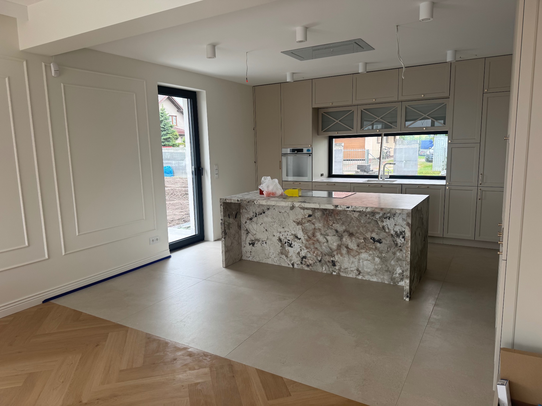 Jasna kuchnia z wyspą z kamienia i parkietem ułożonym w jodełkę. Widoczne wykończenia ścian i zabudowa kuchenna w odcieniach beżu. Oświetlenie sufitowe punktowe.