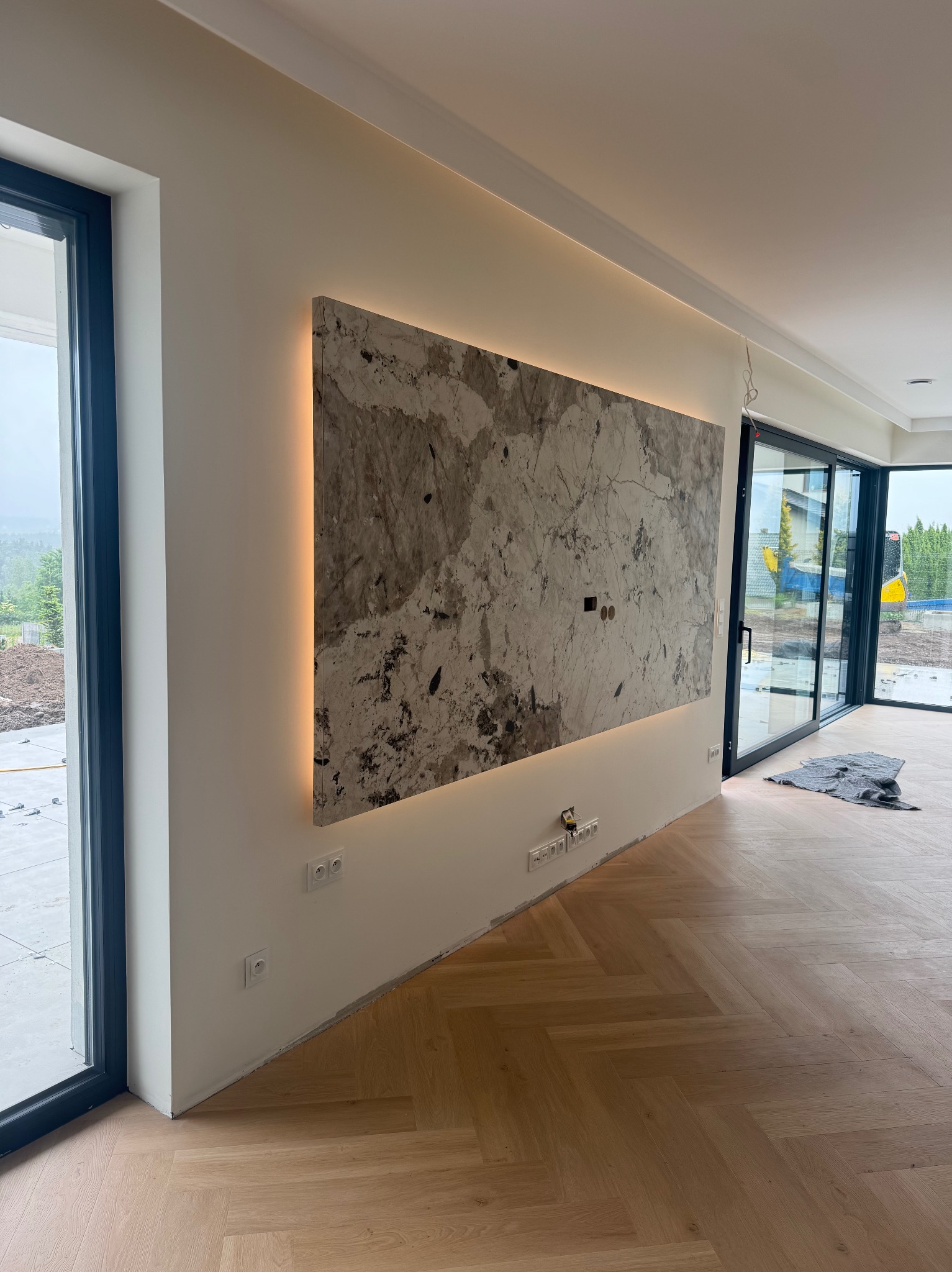 Wykończona ściana z marmurową płytą podświetloną LED, widoczne gniazdka elektryczne i parkiet w jodełkę, duże okna przesuwne z widokiem na zewnątrz.